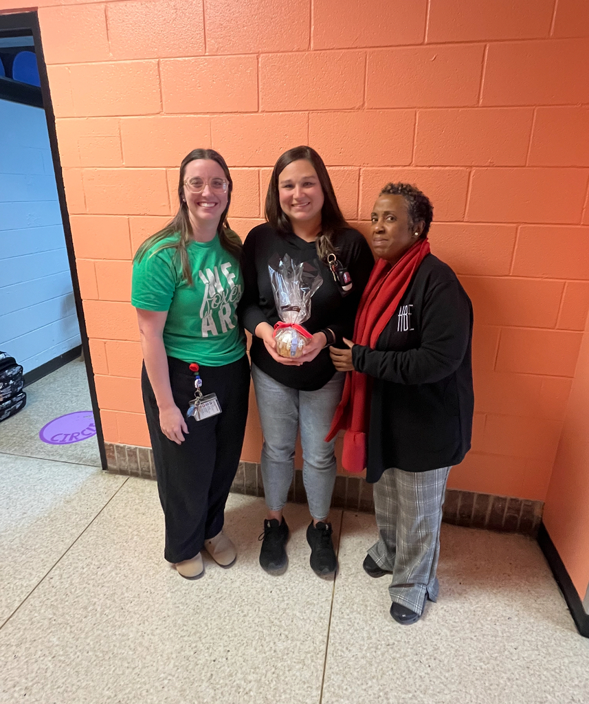 Three adults standing indoors; woman on left wears a green T-shirt reading 'WE LOVE ART', woman on right wears a black sweatshirt reading 'HBE',