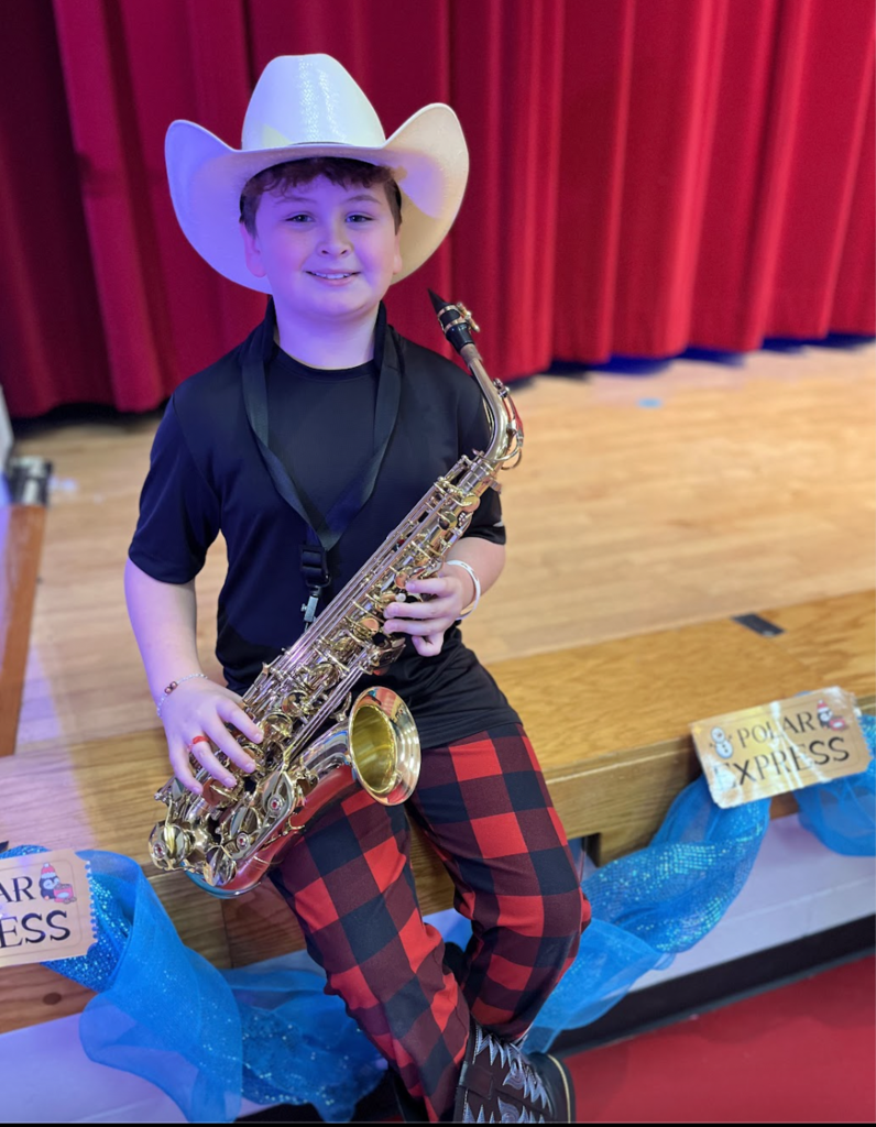 Student sitting on the edge of a stage playing saxophone wearing hats and flannel pants 
