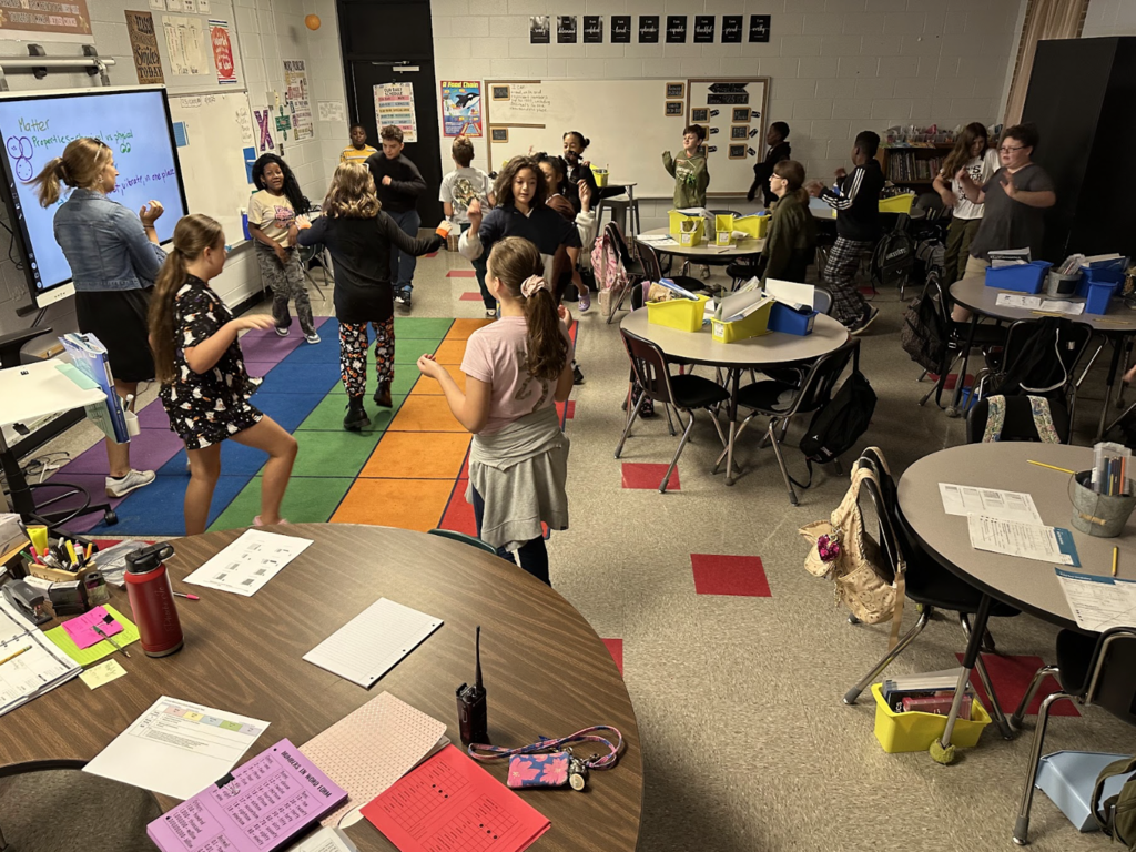 Students moving around their classroom, seen in picture is rainbow carpet and multiple desks,