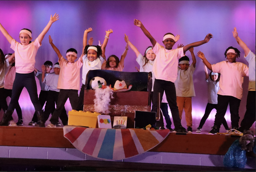 Multiple students standing on stage with arms up over head wearing pink shirts and pink headbands 
