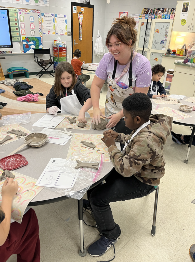 Our art teacher Ms. Deese standing with two students sitting at a desk working on clay.