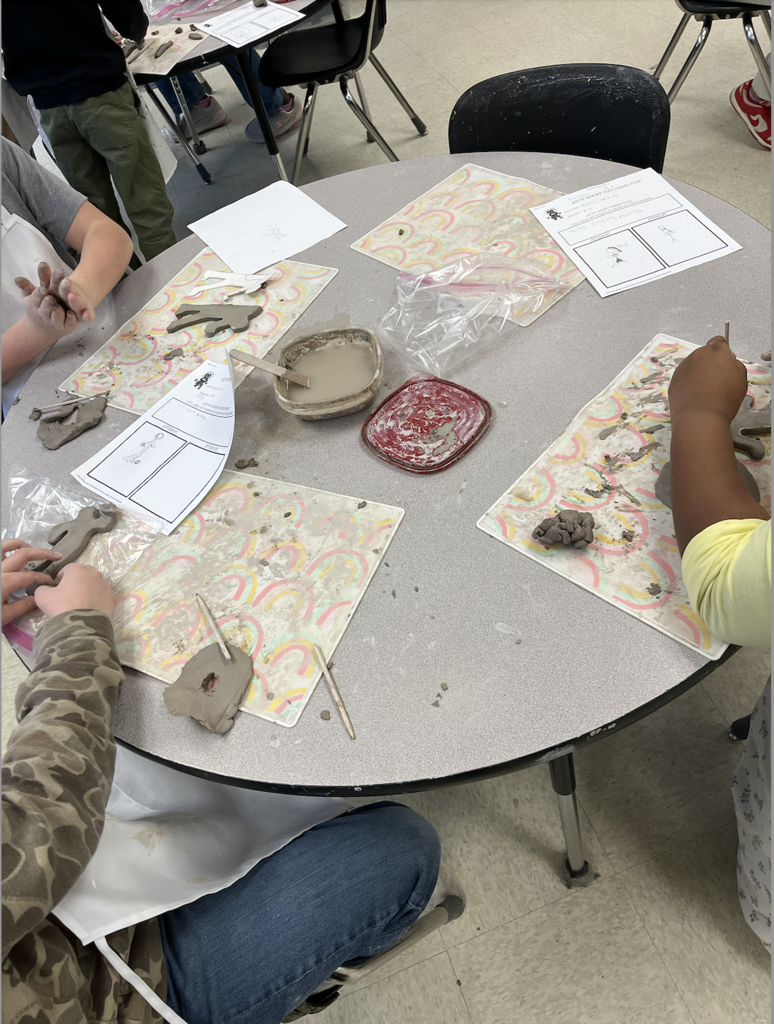 Image shows a desk with three student sitting around it working on clay, There is a bucket of slip in the center.