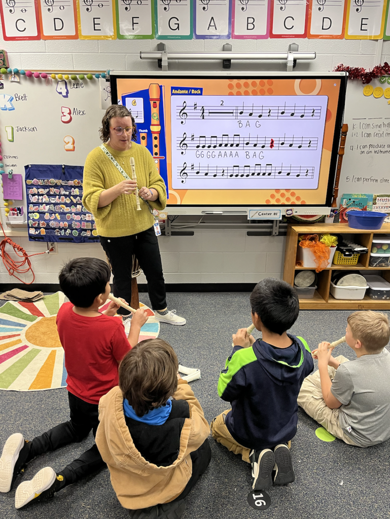 Teacher- Mrs. Turner standing in front of a group of four students playing recorder, on the smartboard behind is music for the students to read and play