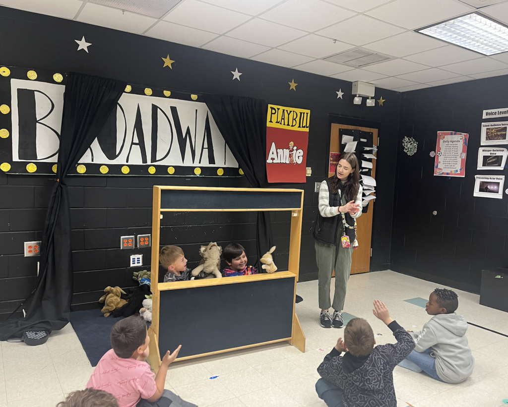 Two students behind the puppet theatre with teacher standing to the right. All puppets, students and teacher are bowing!