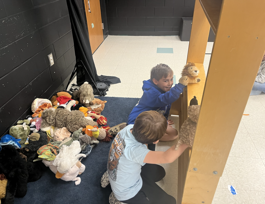 two students crouched behind the puppet theatre holding a lion and an owl with a pile of puppets behind them!