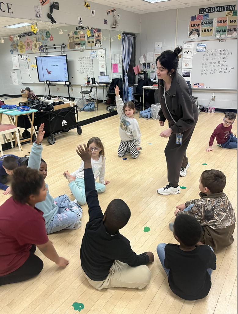 One teacher in green suit learning in talking to students sitting on the floor raising their hand