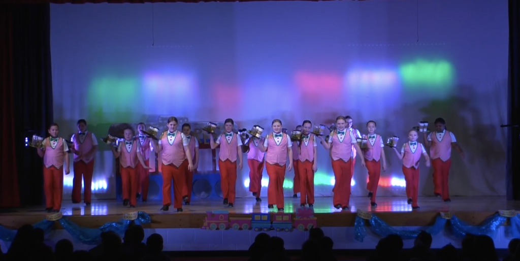 Girls wearing red pants and white and red striped vests holding hot chocolate cups on a tray dancing on stage