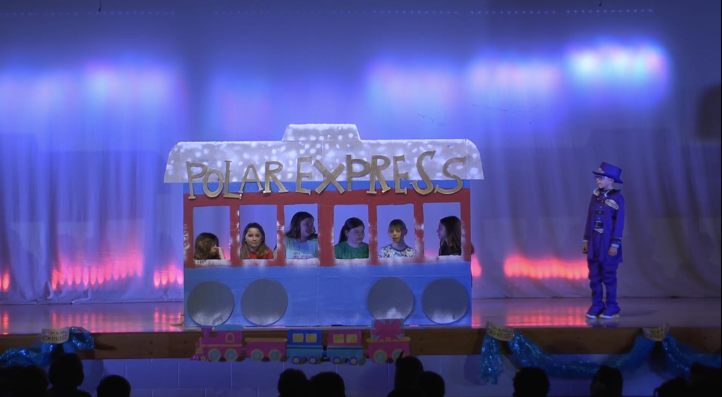 Students on stage sitting in windows of a cut out of the Polar Express with student in purple conductor suit