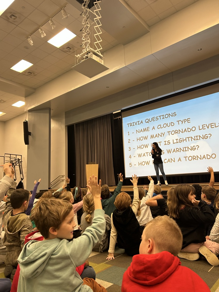 meteorologist presenting on stage with students hands raise 