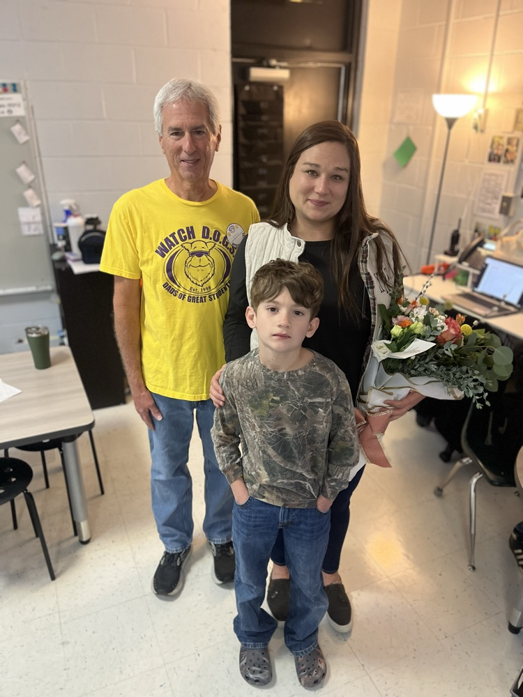Teacher of the Year stands in a classroom holding flowers, with an adult staff member and a young child standing beside her. Classroom tables and technology are visible in the background.
