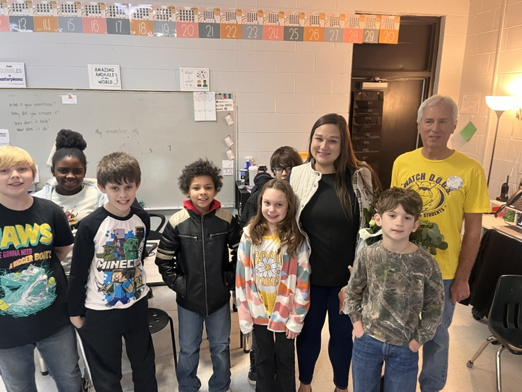 Teacher of the Year stands in a classroom surrounded by smiling elementary students. The teacher holds flowers while students stand in front of desks and a whiteboard.