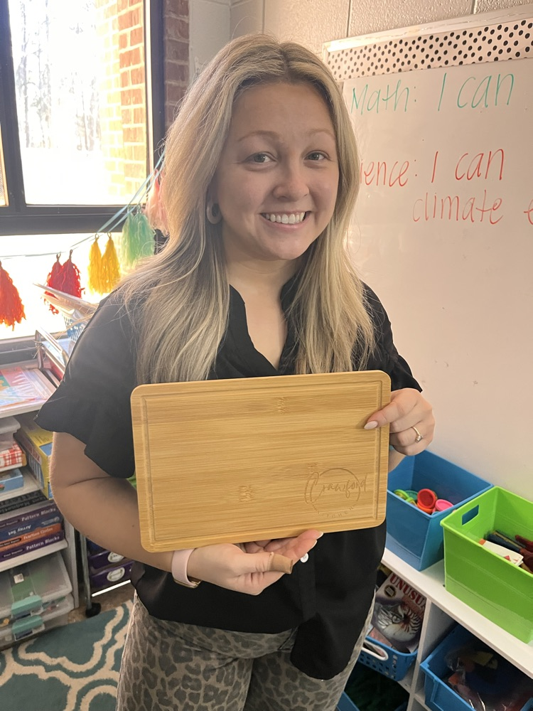 Teacher in black shirt holds cutting board award with name imprinted on it. Standing in a classroom with window and whiteboard directly behind her.  