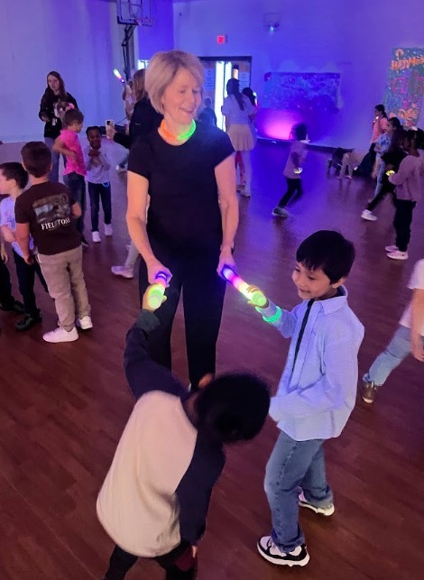 An adult smiles while holding two glowing light sticks and wearing a neon necklace as children nearby reach for glow items on a gym floor with purple lighting.