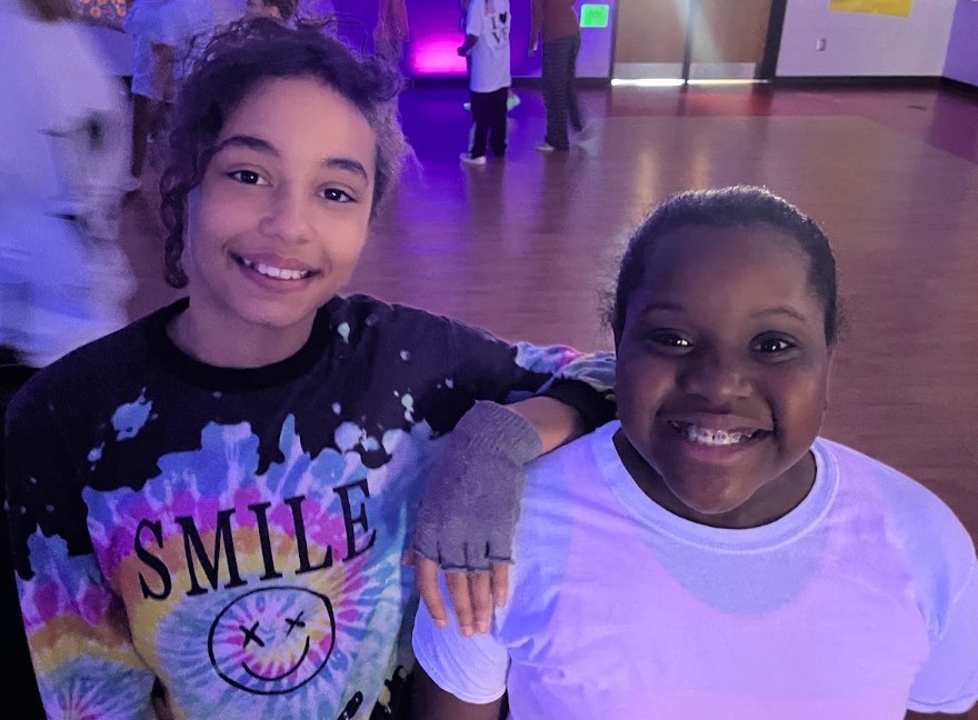 Two girls smile at the camera under purple lighting; the left girl wears a tie‑dye shirt that reads “SMILE :)” and the right girl shows braces.