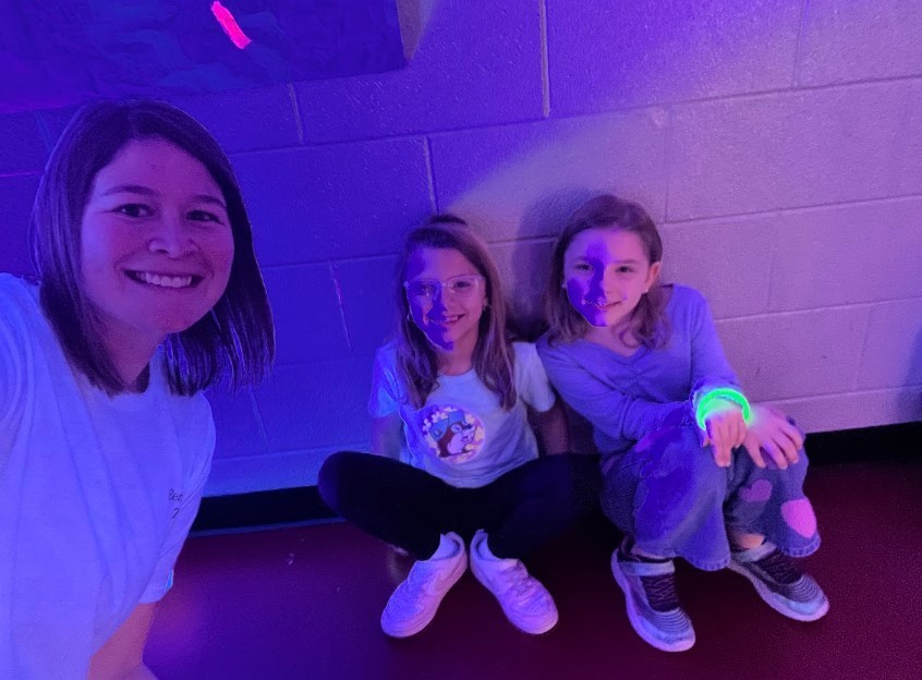 An adult and two girls sit against a brick wall under purple light, all smiling; one girl wears glasses and another has a bright green glowing bracelet.
