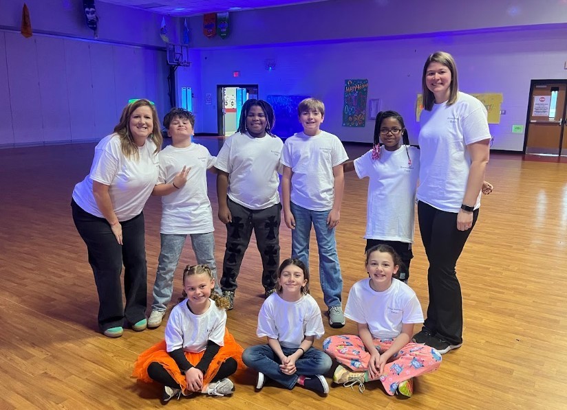 Nine people pose in white shirts and jeans. Shirts are matching, with back row of 6 standing and three down on the floor sitting criss cross.
