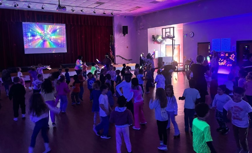 Wide shot of a crowded gym disco with children and adults dancing under purple/blue lights, a stage projection, and a glowing blue display that reads “GLOW UP DISCO.”