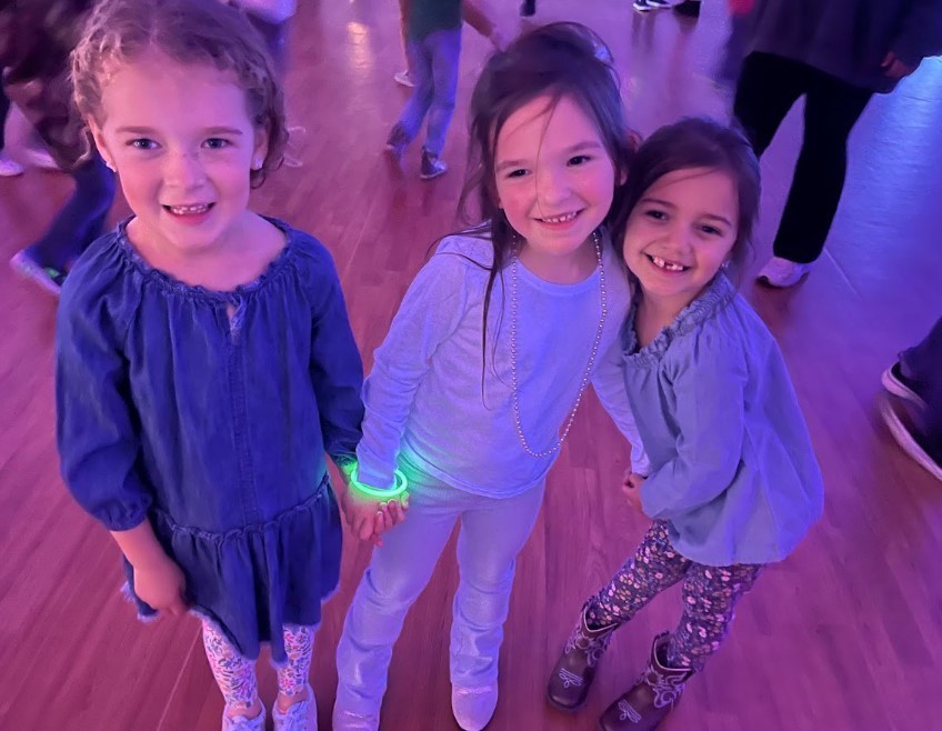 Three girls hold hands and smile at the camera on a wooden floor under purple/pink party lighting; one wears a green glow bracelet and another wears cowboy boots.