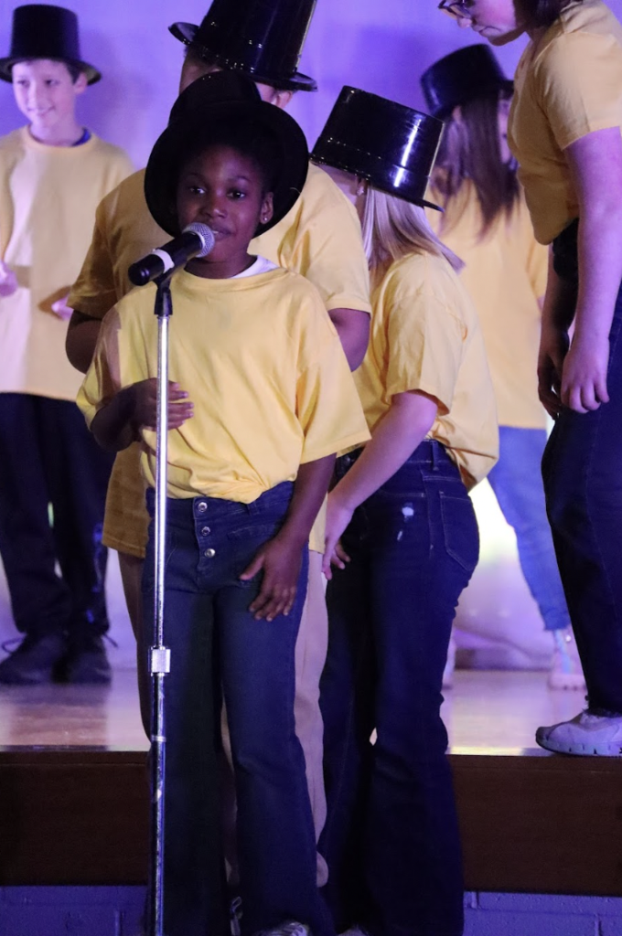 Child wearing yellow shirt standing at microphone on stage