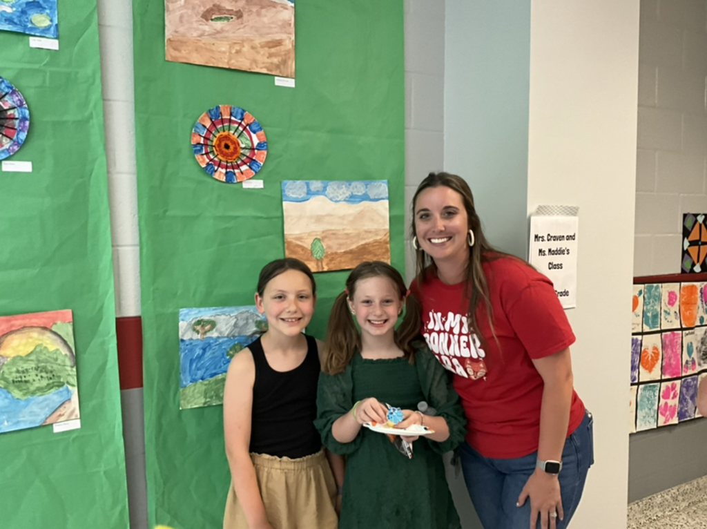 Image of green backdrop with watercolor paintings, two students and one teacher standing in front