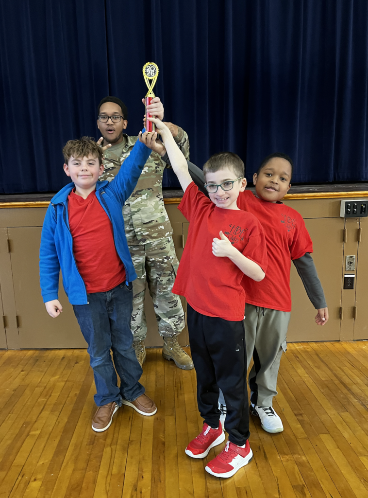 3 students lifiting a trophy into the air with man in uniform in the back