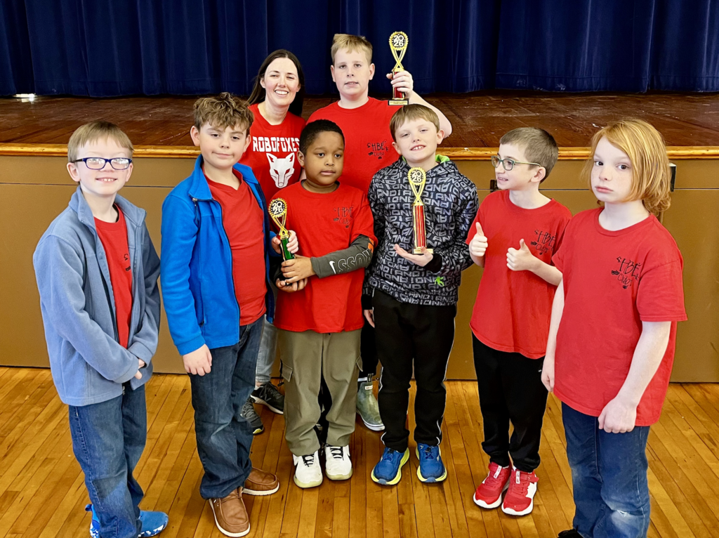 Robotics team, 6 students standing in the front row, one in the center is holding a trophy. One student and one teacher in the second row holding a trophy