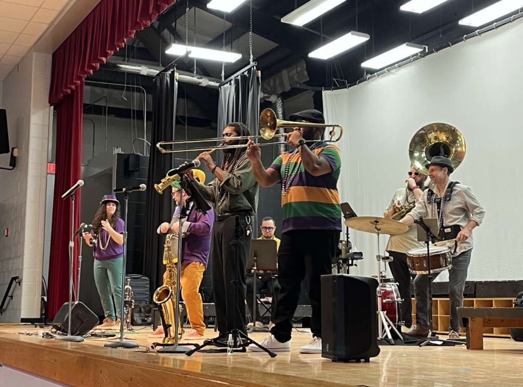 Picture features 7 band members on the stage, wearing gold, purple and green playing snare drum, tuba, trombone, saxophone, tambourine, washboard, and piano.