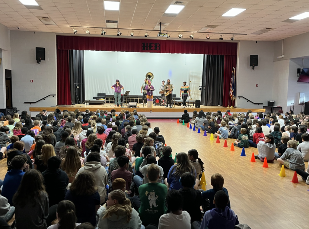 This picture shows multiple (100s) of students sitting on the floor in their MPR facing the stage where a band of 7 people are playing,