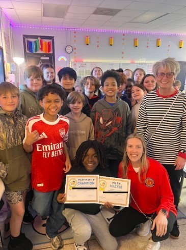 Approximately 15 fifth-grade students and two adult women pose together in a classroom decorated with colorful string lights and pencil cutouts. One student kneels in front holding Math Master Champion and Math Master certificates.
