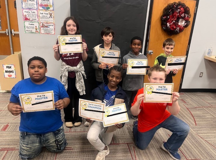 Seven elementary students hold Math Master and Math Master Champion certificates while posing in a classroom. Behind them are educational posters about story genres (Myth, Fairy Tale, Fable, Legend, Folk Tale, Tall Tale) and a Fox Books shelf