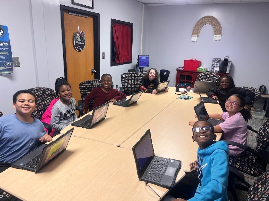 Eight elementary students sit around a large table with laptops in a classroom learning center. They smile at the camera. A decorative macrame rainbow hangs on the back wall, and a red curtain covers a window behind them.