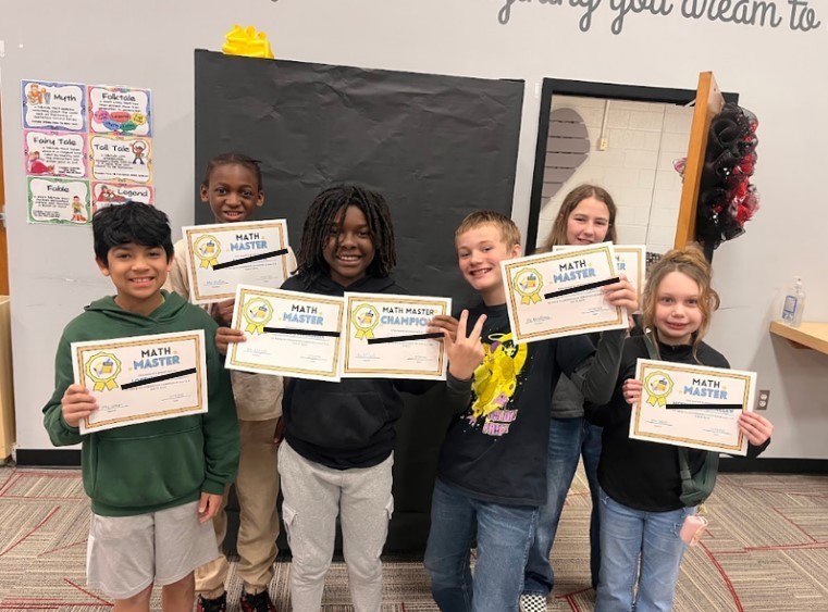 Seven fifth-grade students smile while holding Math Master and Math Master Champion certificates. Behind them are educational posters defining story genres (Myth, Folktale, Fairy Tale, Tall Tale, Fable, Legend) on the classroom wall.