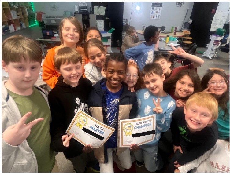 Approximately 14 fourth-grade students pose together in a classroom. Several make peace signs with their fingers. Two students in the center hold Math Master and Math Master Champion certificates.