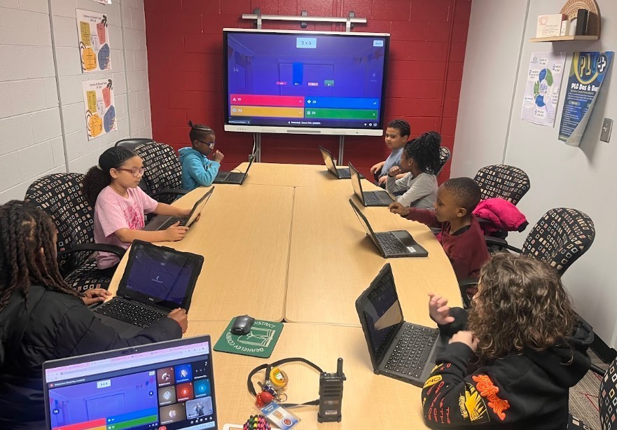 Nine elementary students work on laptops around a table while playing a Kahoot math game. A large screen displays the game showing a multiplication problem (5 x 5) with answer options. Educational posters line the classroom walls.