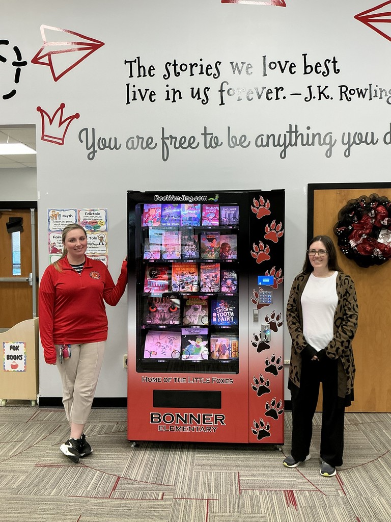 Two educators stand beside a red and black book vending machine at Bonner Elementary, filled with children's books including titles like "Hilda," "The Wild Robot," and "Woodland Whispers." The hallway features inspirational quotes and a "Fox Books" collection box. Paw print decals and genre charts celebrate reading and storytelling.