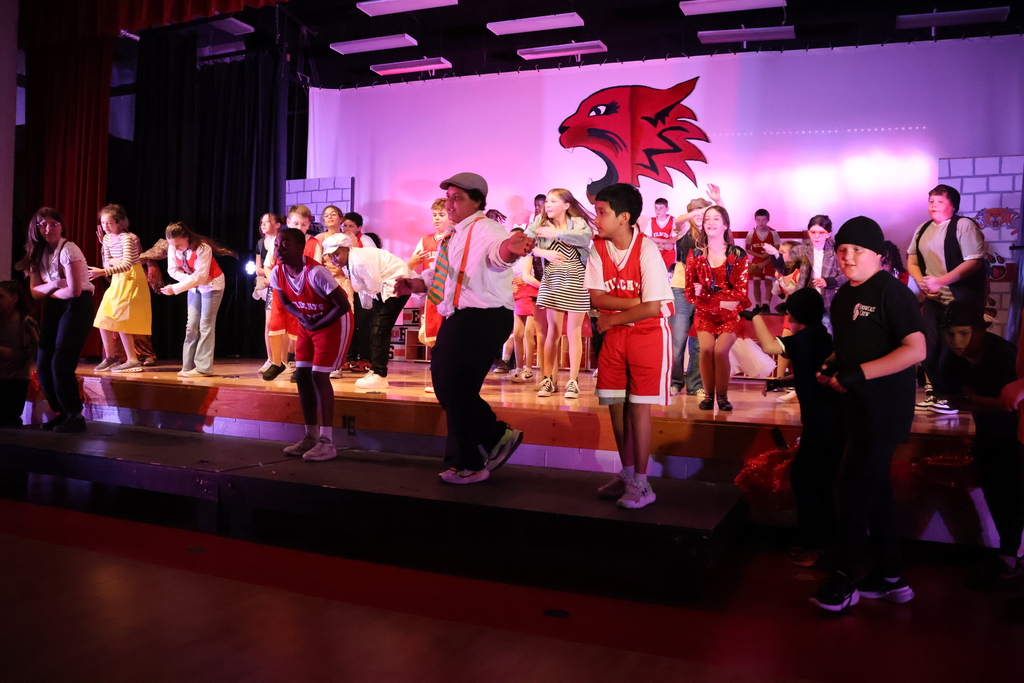 Picture features many fifth grade students dancing on the stage in various costumes from High School Musical with Wildcat Set piece in the background. 