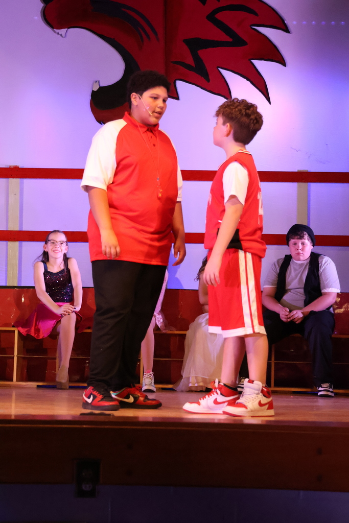 Two students standing centered staged dressed like Coach Bolton and Troy Bolton from high School Musical (basketball jerseys). Facing each other talking.
