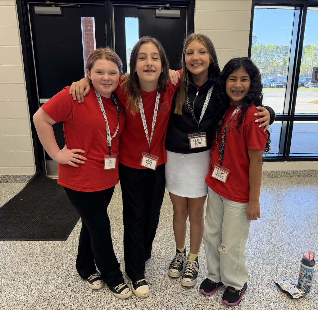 Four students in the lobby of GCHE, wearing red bonner shorts with name tages. All four students have arms around each other and are smiling!