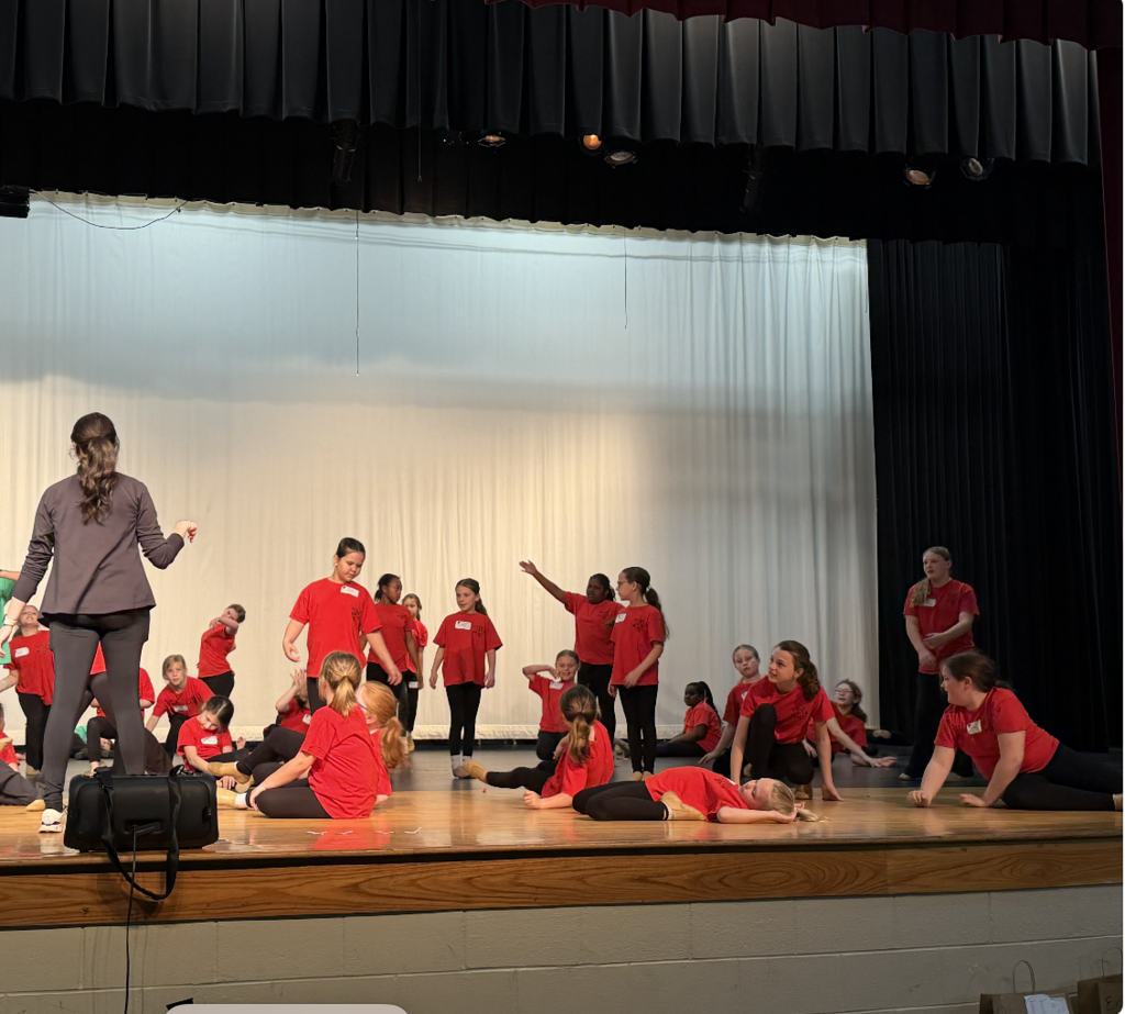 Multiple dancers wearing red shirts dancing on stage with one teacher in the front