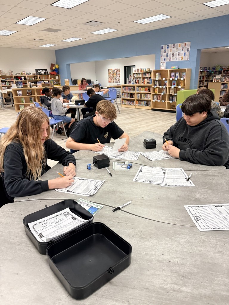 A group of students sit around a table in a library, working on papers with pens.