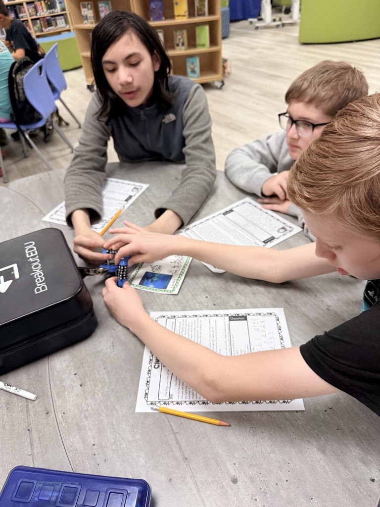 Three children focus on a small project on a table, surrounded by papers and pencils.