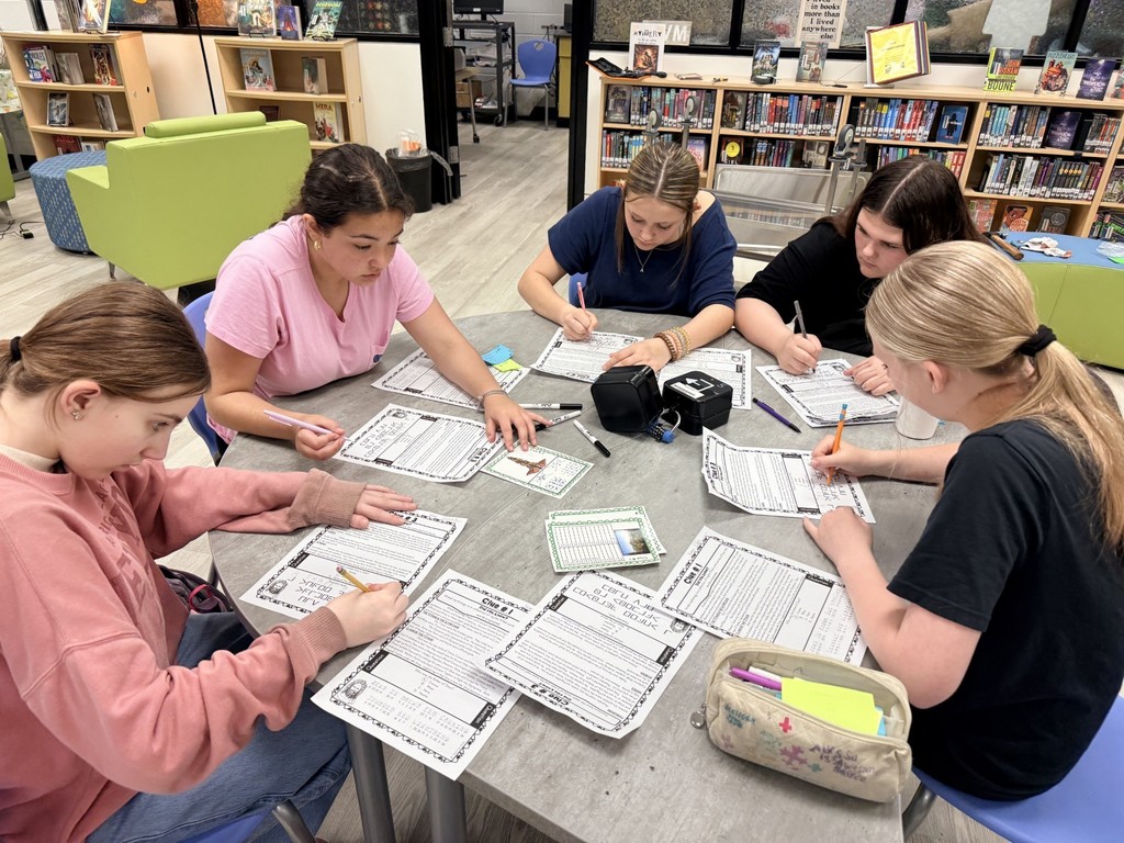 Five Students seated around a table in a library, each holding a pen and working on papers.