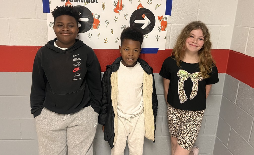Three students standing in a school hallway in front of educational posters about breakfast and nutrition.