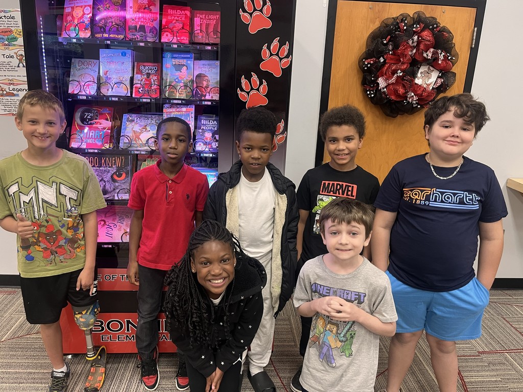 Group of seven elementary school children posing in front of a book vending machine displaying children's book titles, celebrating their achievements.