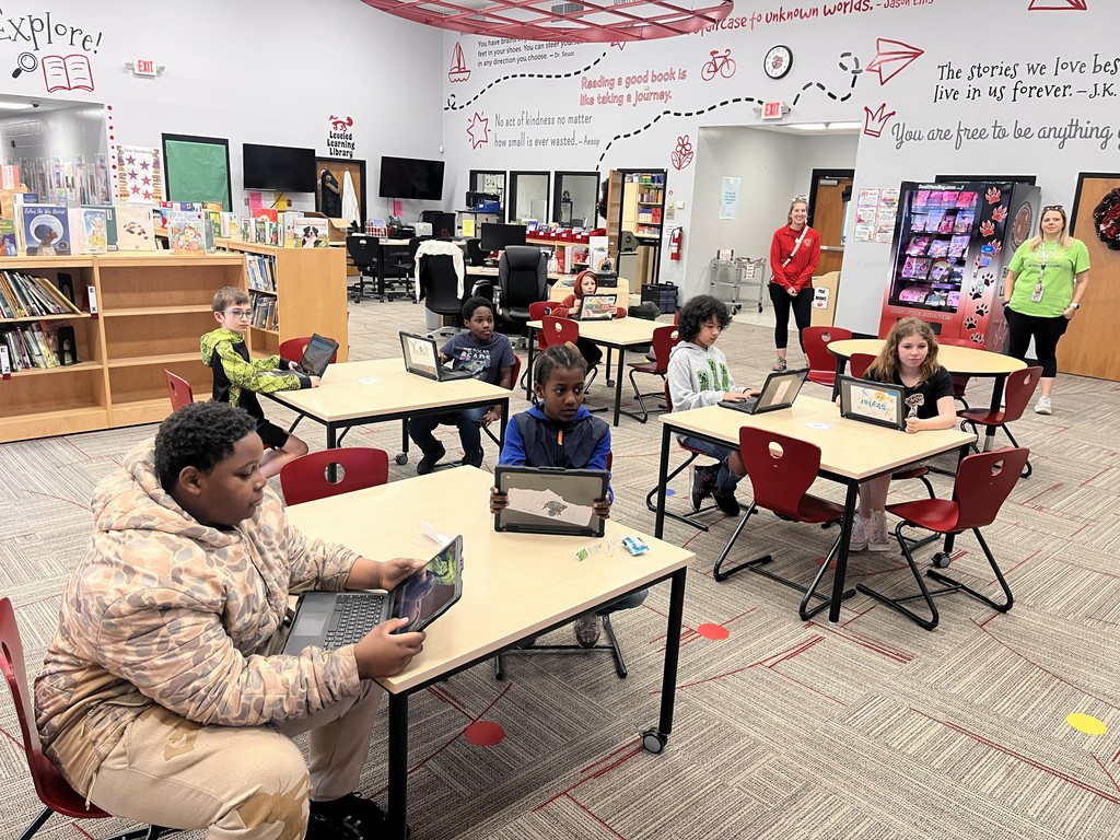 Students working on laptops at tables in a modern school library learning center with inspirational quotes on the walls and a book vending machine.
