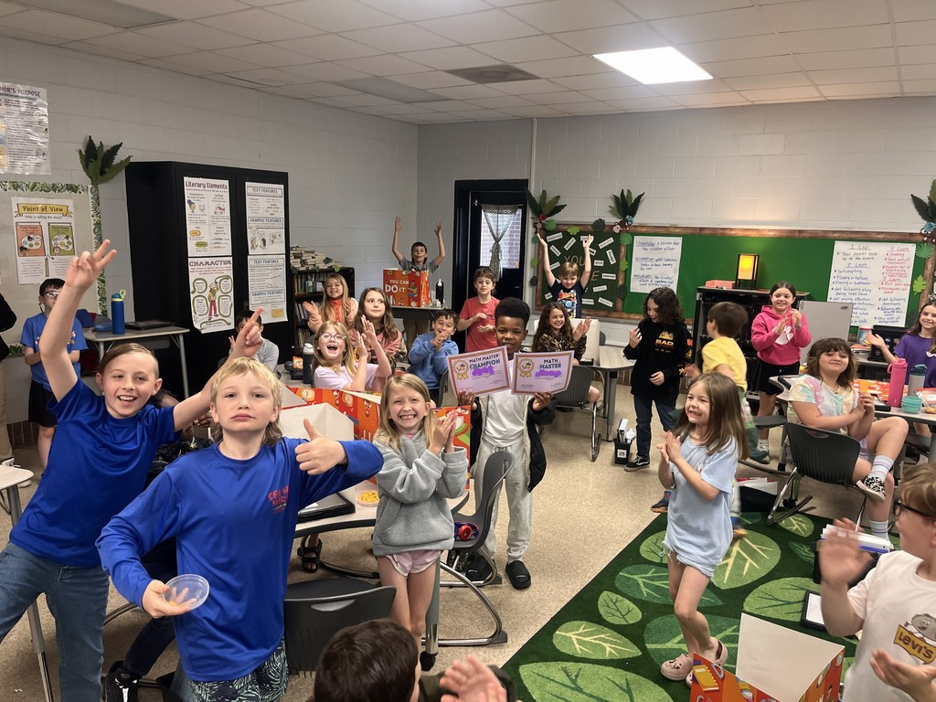 A classroom filled with children. Some children are raising their hands and holding objects. Others are seated at tables.