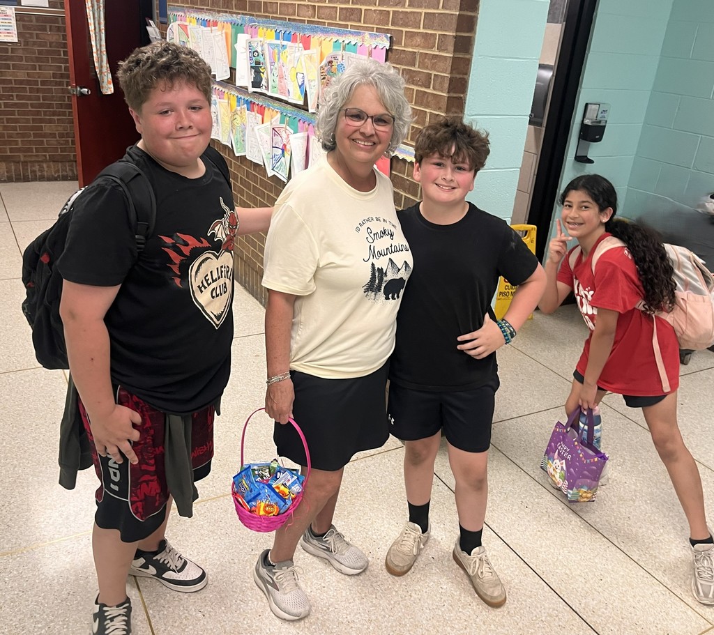 A woman stands with three children, holding baskets. The children wear backpacks and shorts. Behind them, a wall displays colorful papers and a brick wall.