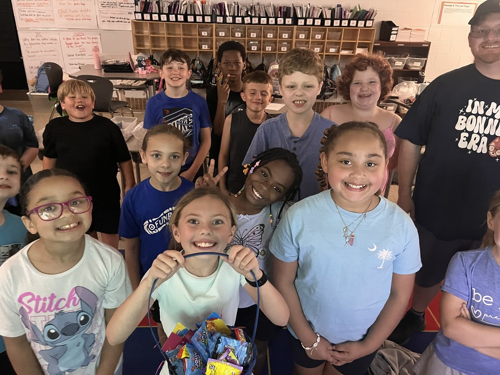 Group of smiling children in a classroom, standing and holding a gift bag. One child wears glasses and a watch.