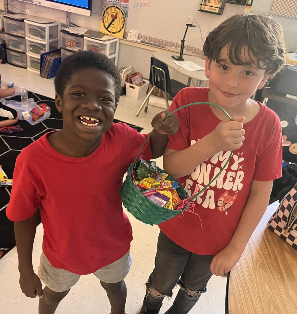 Two boys stand close, one holding a candy basket, both smiling. Behind them is a classroom with a rug, chairs, and shelves.