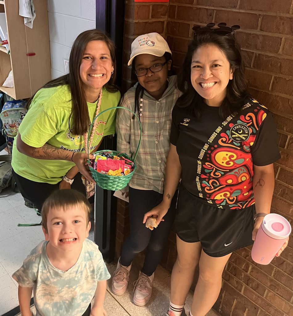 Four people smile next to a brick wall. Two women wear green and black shirts; one holds a basket. A boy wears a tie-dye shirt.
